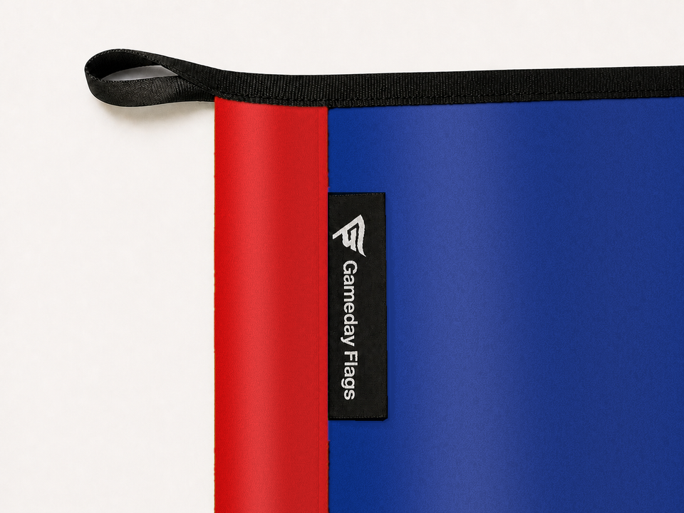 Buffalo Bills Authentic Gameday Flag construction detail — restrained woven-satin Buffalo Bills Accent sewn header, textured Buffalo Bills Primary face, crisp woven Gameday Flags tag and controlled contact shadow, black loop, perimeter webbing, and stitching — source_sha256=2dacc15d6f768aa2fedbb8582ba8daf74754fc39751eac33d91b590a854f1beb