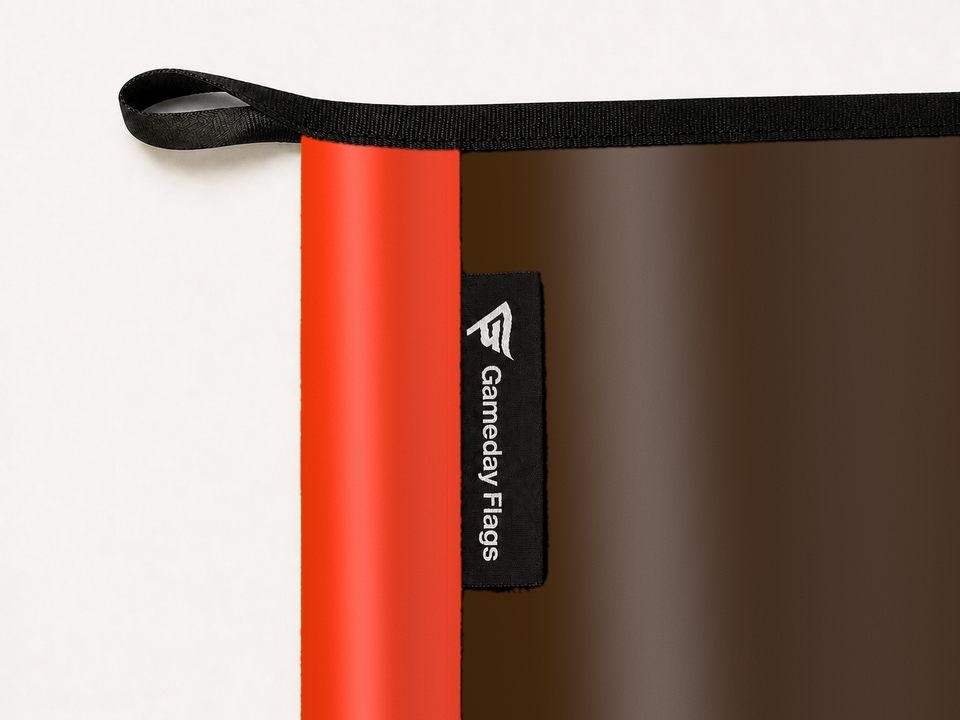 Cleveland Browns Authentic Gameday Flag construction detail — restrained woven-satin Cleveland Browns Accent sewn header, textured Cleveland Browns Primary face, woven Gameday Flags tag, black loop, perimeter webbing, and stitching — source_sha256=73b462eaa4721198ce6dc852c92cce703bd1e8a8afc30f3ab299309b4246ff26