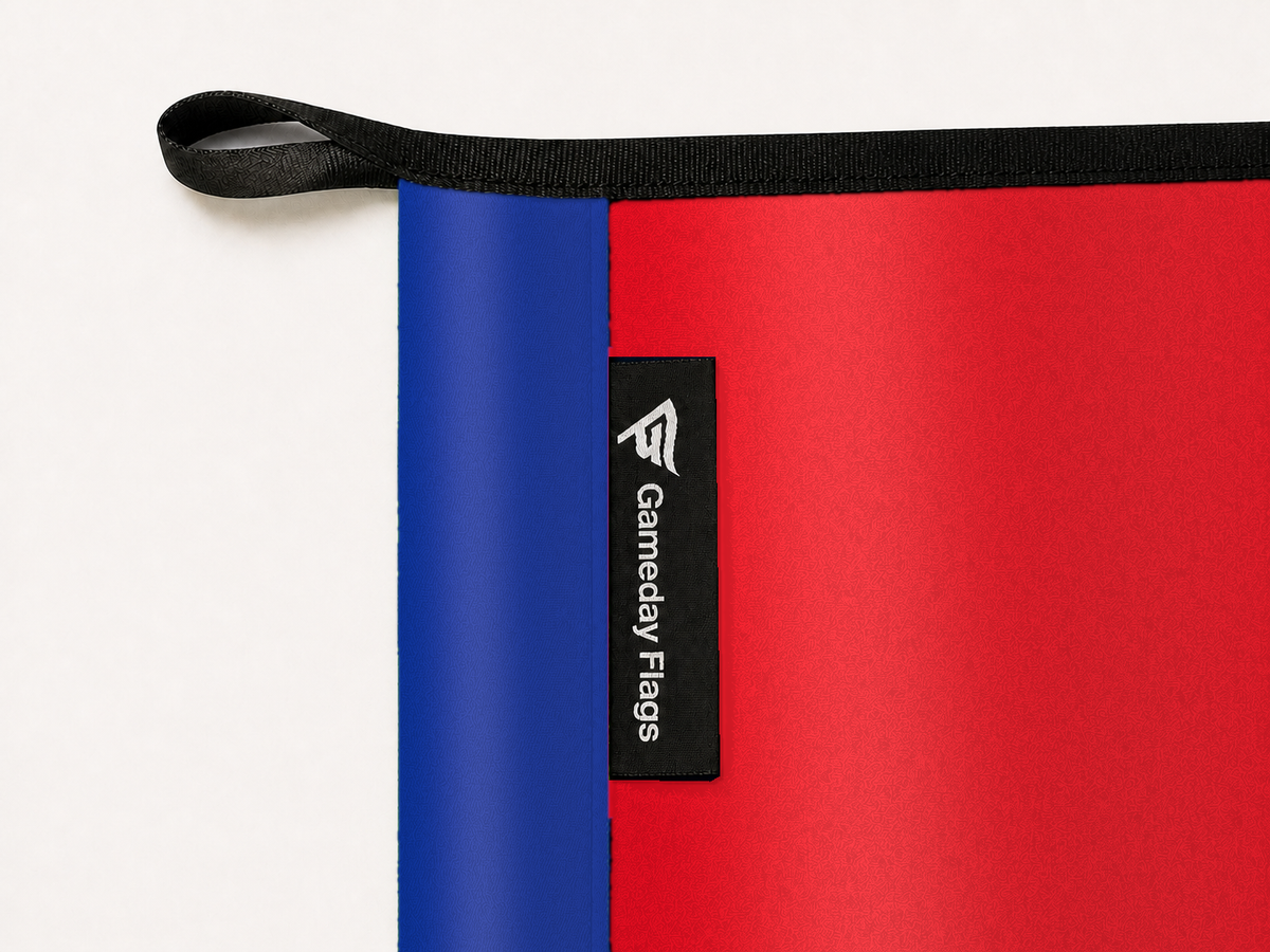 Arsenal Authentic Gameday Flag construction detail — restrained woven-satin Arsenal Accent sewn header, textured Arsenal Primary face, crisp woven Gameday Flags tag and controlled contact shadow, black loop, perimeter webbing, and stitching — source_sha256=ea0762de6cd23d02853a82fb0ff99e4ef40a8a2e4a926d12aff12e8a9e9e9f4f