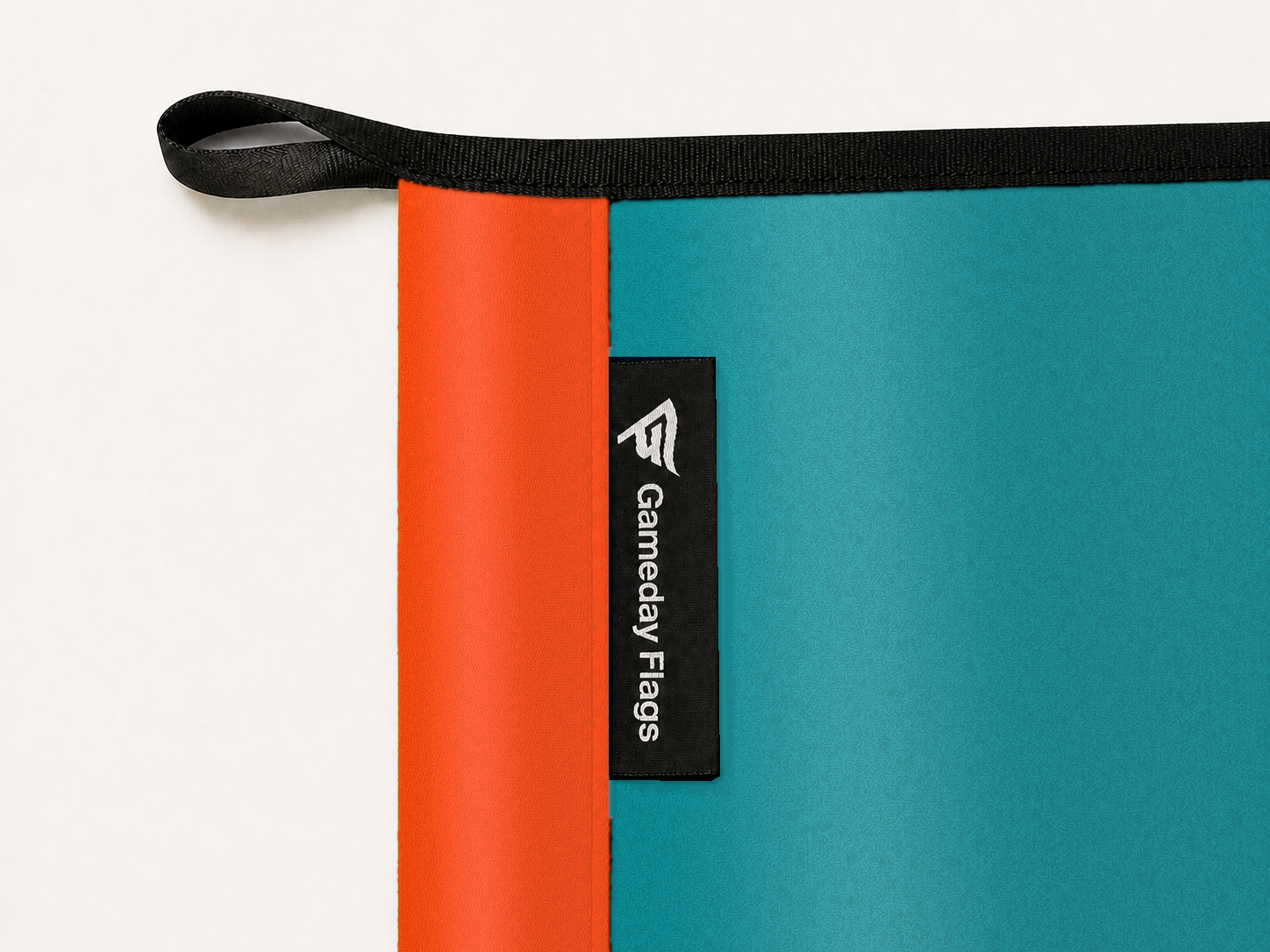 Miami Dolphins Authentic Gameday Flag construction detail — restrained woven-satin Dolphins Orange sewn header, textured Dolphins Aqua face, crisp woven Gameday Flags tag and controlled contact shadow, black loop, perimeter webbing, and stitching — source_sha256=fc306918528a336dc1984a8e926200d867a8c5d4686c5019b7af2832ba6e3ee2