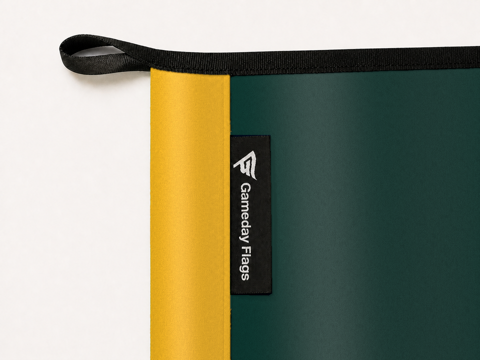Athletics Authentic Gameday Flag construction detail — restrained woven-satin Athletics Accent sewn header, textured Athletics Primary face, crisp woven Gameday Flags tag and controlled contact shadow, black loop, perimeter webbing, and stitching — source_sha256=4c7558fcd1456a5dc3ef167579176c615be756fcfd6d1d99e6bdaa7d684fd8d0