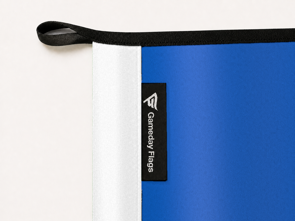 Buffalo Bulls Authentic Gameday Flag construction detail — restrained woven-satin Buffalo Bulls Accent sewn header, textured Buffalo Bulls Primary face, crisp woven Gameday Flags tag and controlled contact shadow, black loop, perimeter webbing, and stitching — source_sha256=7c4cfd71e27aa5ee086b77e9b7044fed132db6ebf883f607872f31ae1a76a7fd