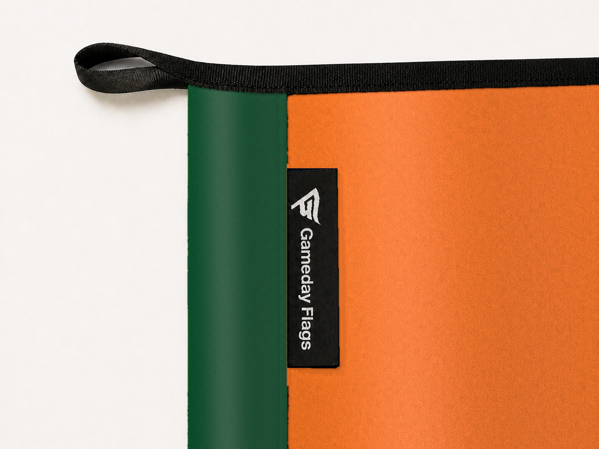 Miami Hurricanes Authentic Gameday Flag construction detail — restrained woven-satin Miami Hurricanes Accent sewn header, textured Miami Hurricanes Primary face, crisp woven Gameday Flags tag and controlled contact shadow, black loop, perimeter webbing, and stitching — source_sha256=74c50d84c20e92aa282e5846d5232137dec9d8211b929b824d84f6e8108c1f7f