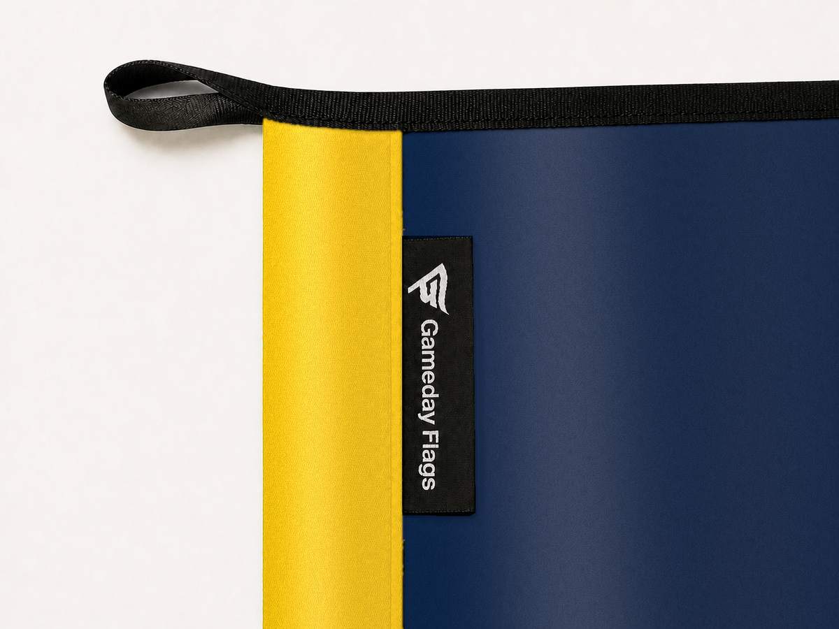 Michigan Wolverines Authentic Gameday Flag construction detail — restrained woven-satin Michigan Wolverines Accent sewn header, textured Michigan Wolverines Primary face, crisp woven Gameday Flags tag and controlled contact shadow, black loop, perimeter webbing, and stitching — source_sha256=b139d6f18c44b051a9451e5d0478bd01c0ffd3292dea507002bc2b62c71deace