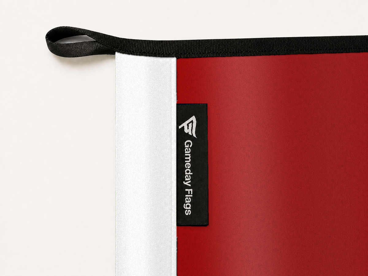 Wisconsin Badgers Authentic Gameday Flag construction detail — restrained woven-satin Wisconsin Badgers Accent sewn header, textured Wisconsin Badgers Primary face, crisp woven Gameday Flags tag and controlled contact shadow, black loop, perimeter webbing, and stitching — source_sha256=2aab1a76190d04c4ec11a188ef4905f5afaf5237a169efcd101b69693471b442
