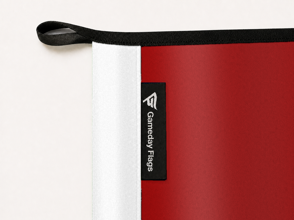 Wisconsin Badgers Authentic Gameday Flag construction detail — restrained woven-satin Wisconsin Badgers Accent sewn header, textured Wisconsin Badgers Primary face, crisp woven Gameday Flags tag and controlled contact shadow, black loop, perimeter webbing, and stitching — source_sha256=2aab1a76190d04c4ec11a188ef4905f5afaf5237a169efcd101b69693471b442